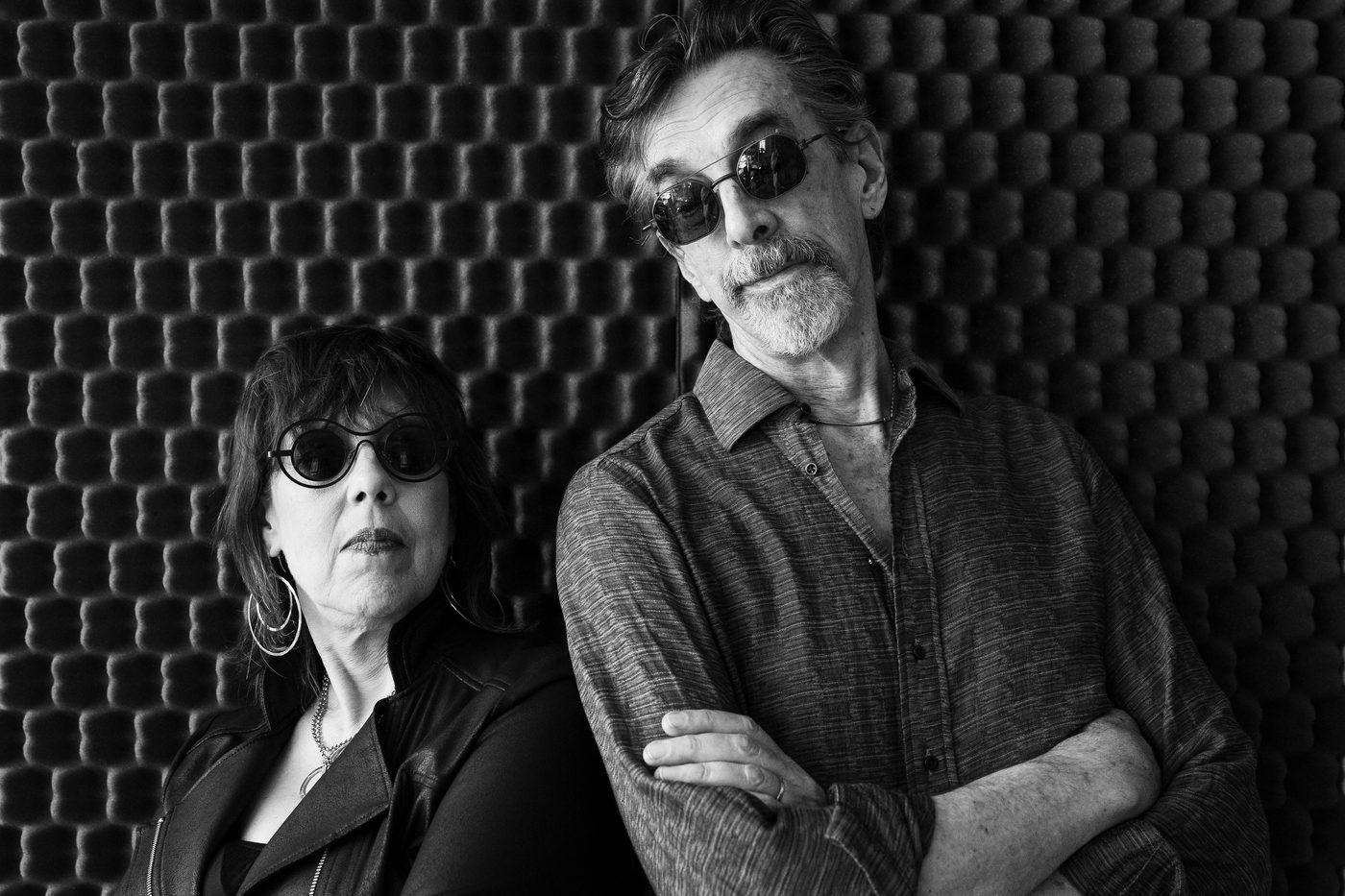 Beth Falcone and Ritt Henn in sunglasses against a studio foam wall, in black and white.