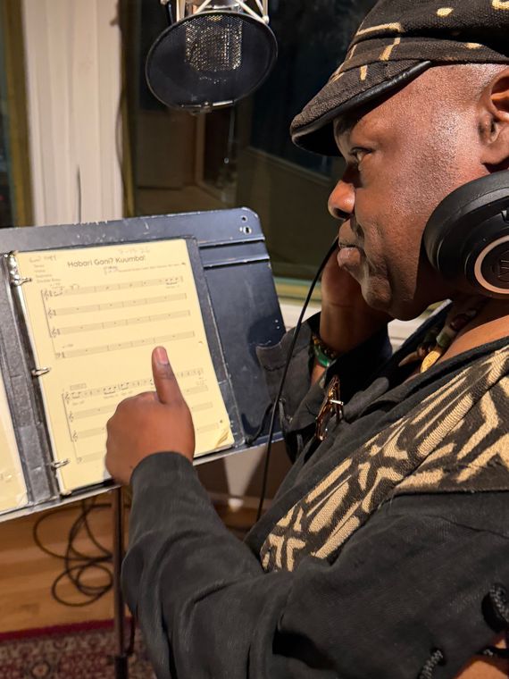 A vocalist in headphones points to the score of Habari Gani? Kuumba! in the vocal booth.