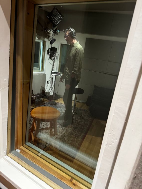 A singer in headphones in the vocal booth, seen through the glass.