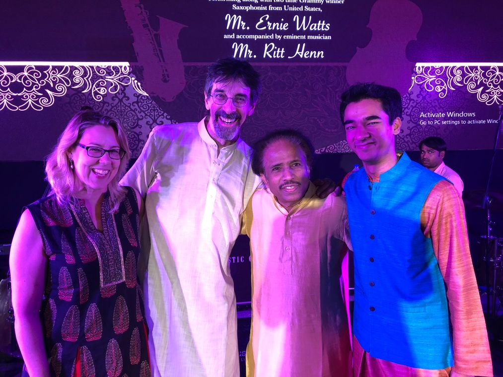 Four musicians on stage in India, beneath a screen announcing saxophonist Ernie Watts, &ldquo;accompanied by eminent musician Mr. Ritt Henn.&rdquo;